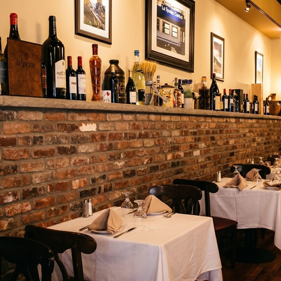 Cozy Italian restaurant interior with brick walls, wine bottles on a shelf, and set tables.