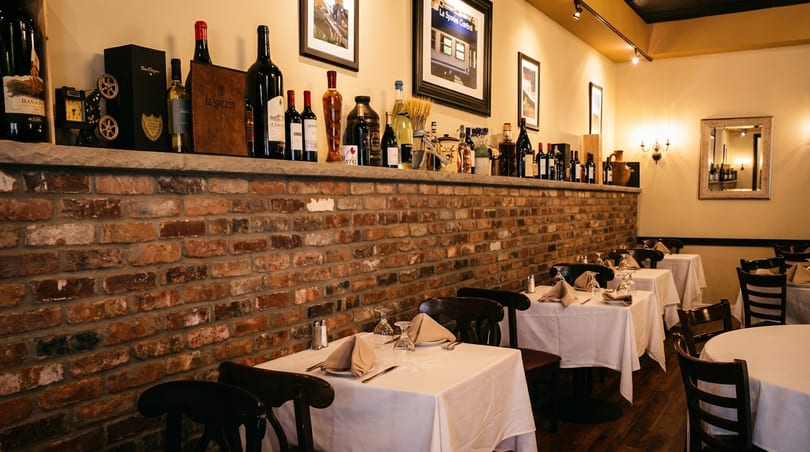 Cozy Italian restaurant interior with brick walls, wine bottles on a shelf, and set tables.