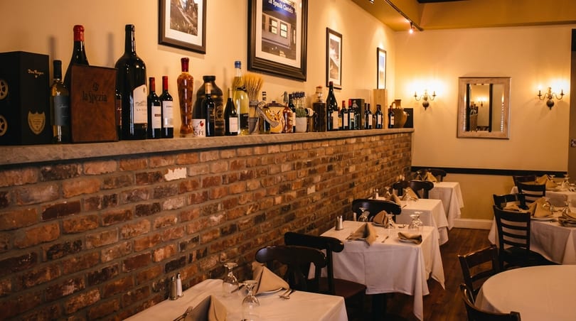 Cozy Italian restaurant interior with brick walls, wine bottles on a shelf, and set tables.